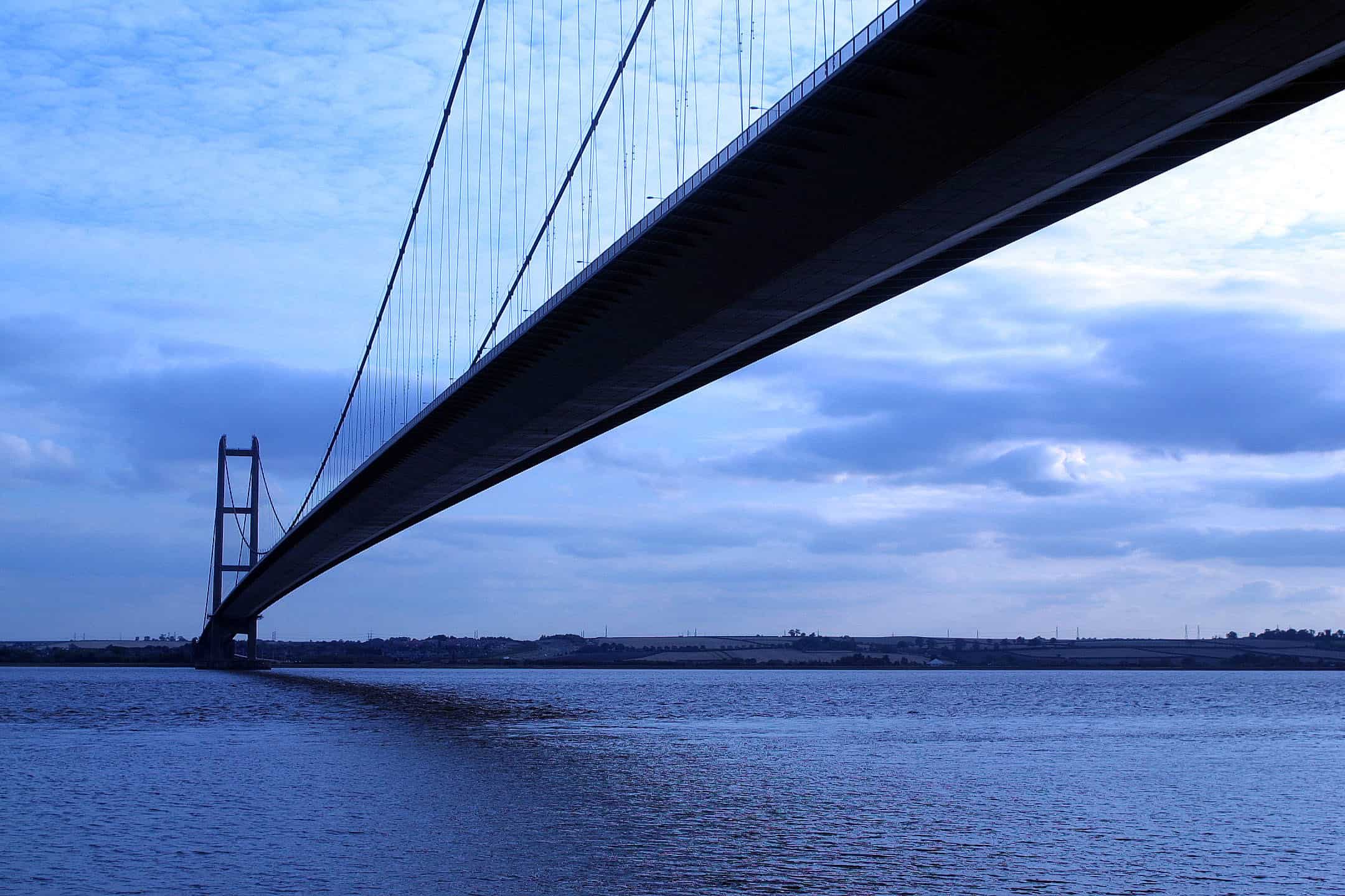 An image of the Humber Bridge, taken at sunrise from below the bridge on the Hull side of the estuary. The fields behind the South Bank can be seen in the distance, on the other side of the water.