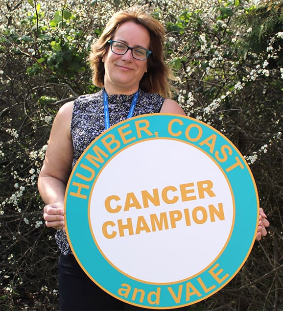 Emma, Volunteer Co-ordinator for the Cancer Champion Programme Humber, Coast and Vale Cancer Alliance, holding a sign advertising the Cancer Champion training. She's standing in front of a green bush with white flowers.