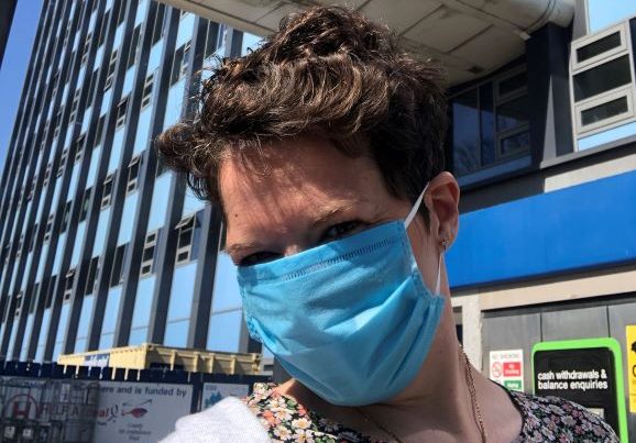 This photo is of Lizzie stood outside Hull University Teaching Hospitals Trust main tower block. There are windows surrounded by blue cladding and Lizzie is wearing a blue facemask, floral t-shirt and grey cardigan. She has short brown hair.
