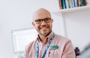 Image of Dr Dan Cottingham sat in a GP office wearing a pink shirt, NHS lanyard and Macmillan name badge. Dan is wearing glasses and smiling. There is a computer screen and book shelf behind him.