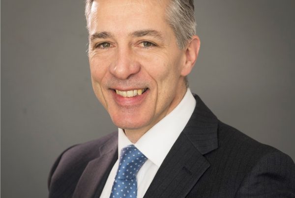 A headshot of Dr Nigel Wells, Clinical Lead for the Humber, Coast and Vale Health and Care Partnership. He's smiling and wearing a black suit jacket over a wh