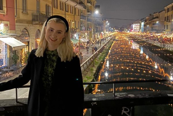 Beckie, a girl in her mid-20s, standing in front of a canal in Prague, smiling at the camera. It's night time and the canal is lit up.