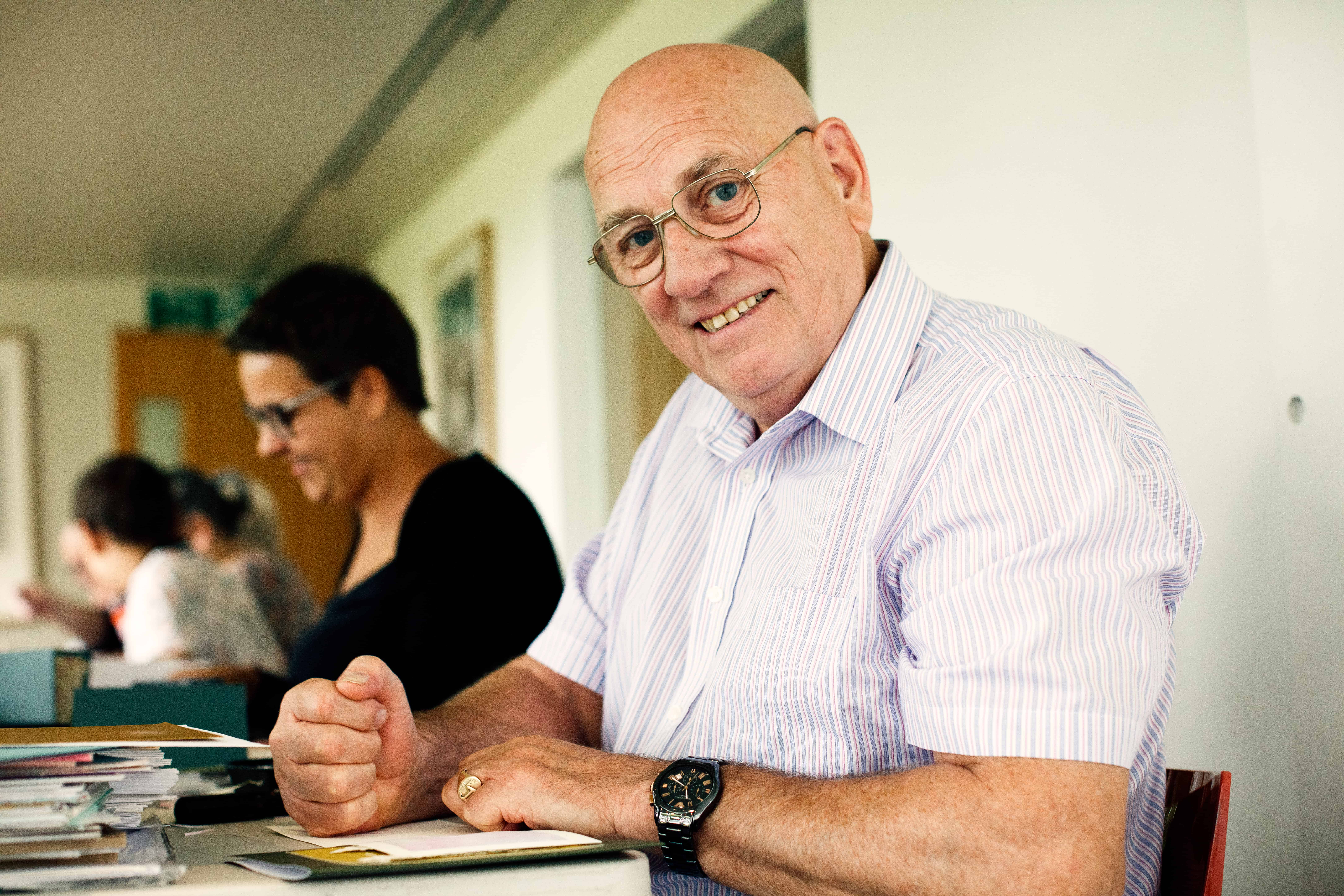 Photo of a man looking at the camera and smiling. The man is white, has no hair and is wearing glasses. He is sat a table with a pen in his hand. There are papers on the table and he is sat next to four other people.