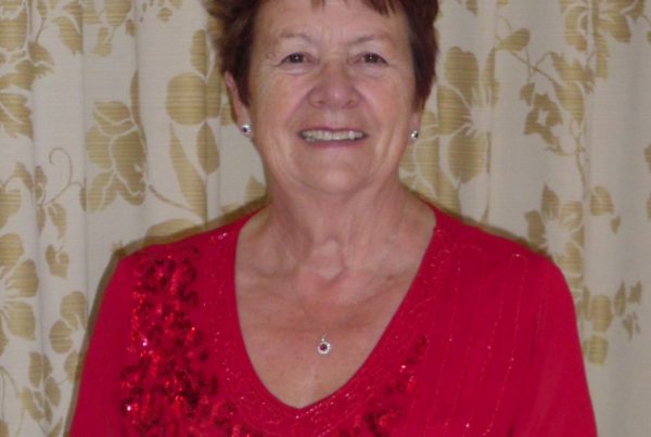 Christine, a pancreatic cancer survivor, looks into the camera and smiles. She is wearing a red sparkly long-sleeved top and has brown cropped hair.