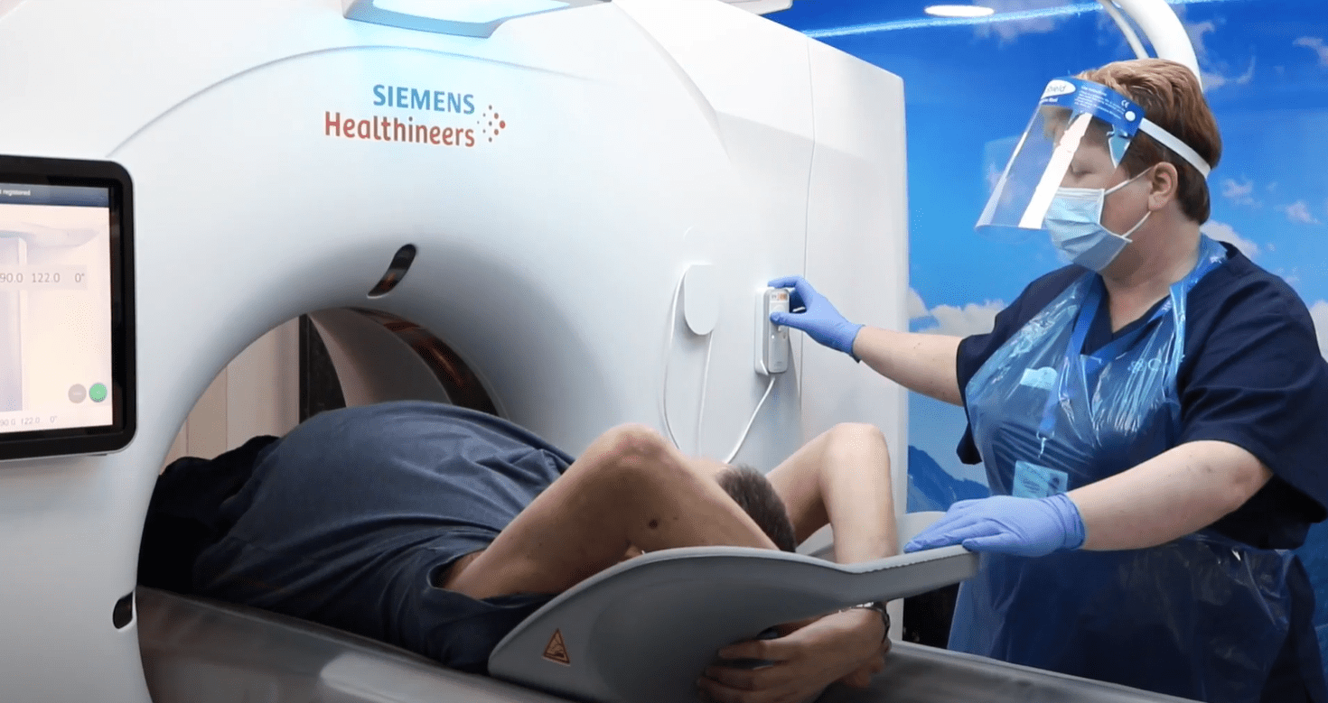 Image of a patient receiving a CT scan as part of a lung health check. The radiographer is wearing a face visor, apron and gloves.