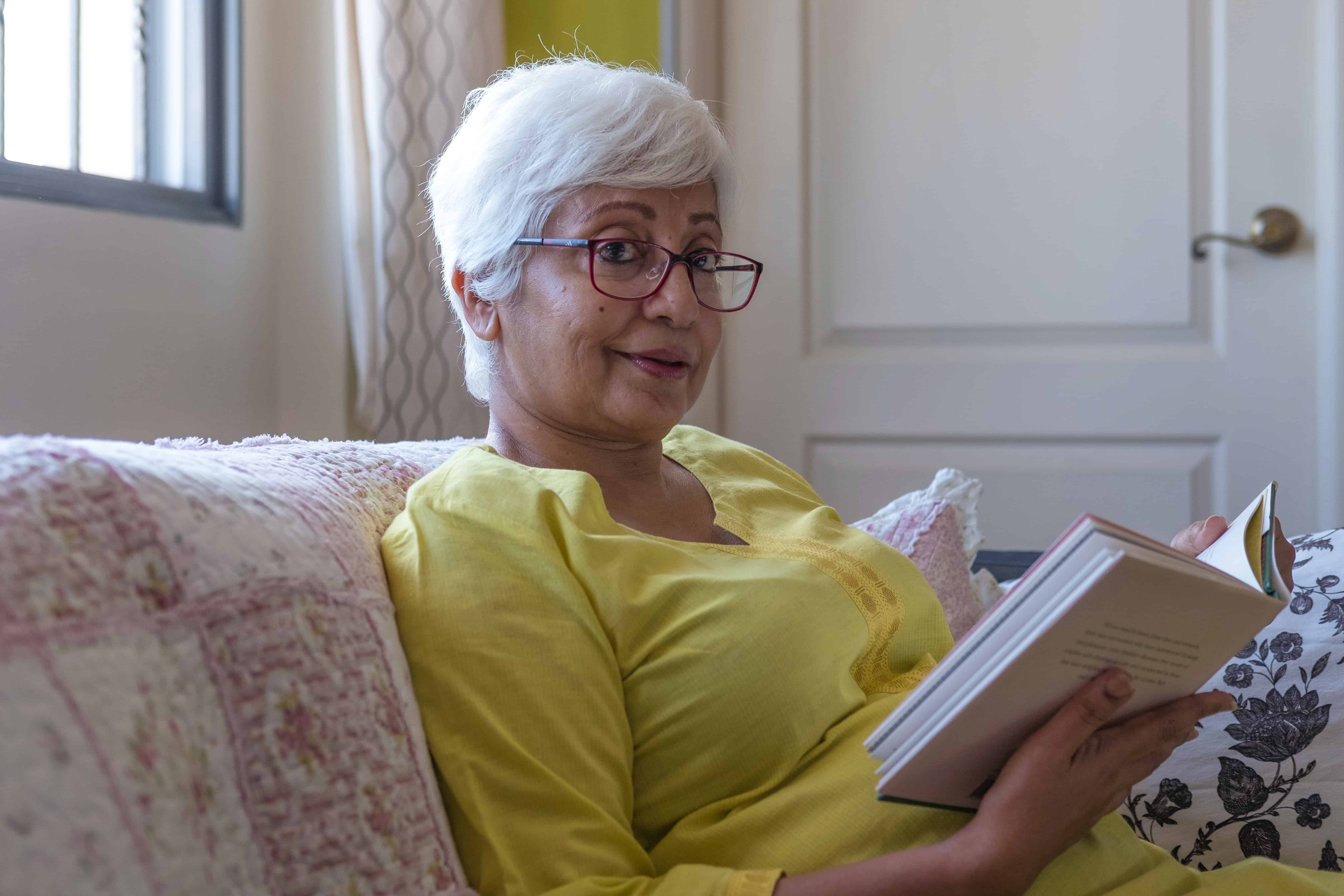 An older woman holding a book, She is sitting on a sofa, wearing a yellow long-sleeved t shirt while looking into the camera. The image is used to represent a cancer patient who has completed the Cancer Quality of Life Survey.