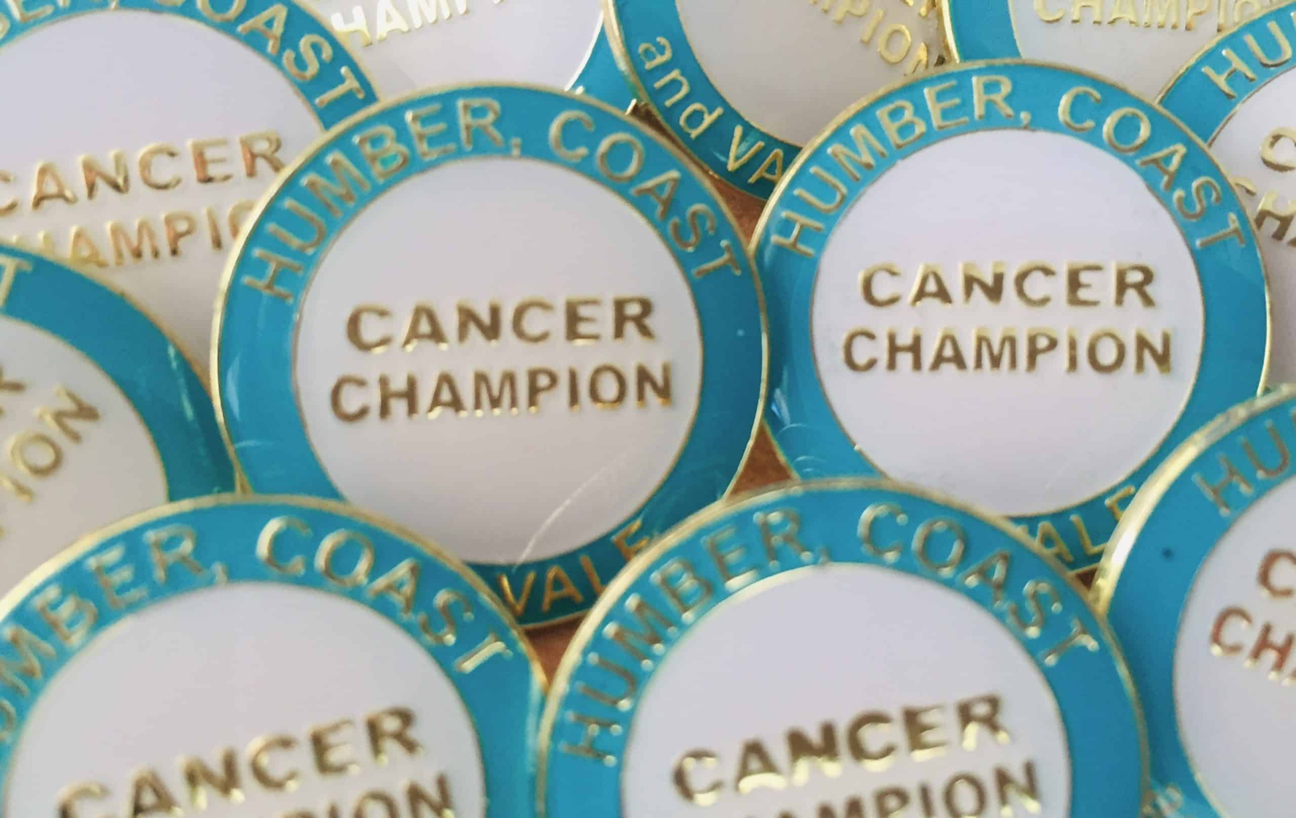 Up close photo of Humber, Coast and Vale Cancer Alliance Cancer Champion badges. The badge is a white circle with a blue border, and they say 'Cancer Champion' in the middle.