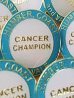 Up close photo of Humber, Coast and Vale Cancer Alliance Cancer Champion badges. The badge is a white circle with a blue border, and they say 'Cancer Champion' in the middle.